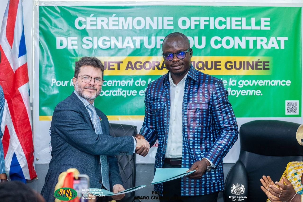 Poignée de main entre le CNOSCG et l'Ambassade du Royaume-Uni devant la banderole du projet Agora Civique Guinée